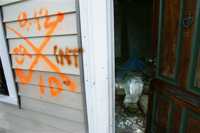 A shrouded body lies on a bed inside a home with spray paint markings indicating its presence in the Mid City section of New Orleans. The number at the top, 9.12, refers to the date that the house was searched. The letters at the left, CA, signify that the search team was from California. The number and letter at the bottom, 1D, stand for one dead person found inside the house. The letters at right, INT, mean the property was entered (refers to interior). 